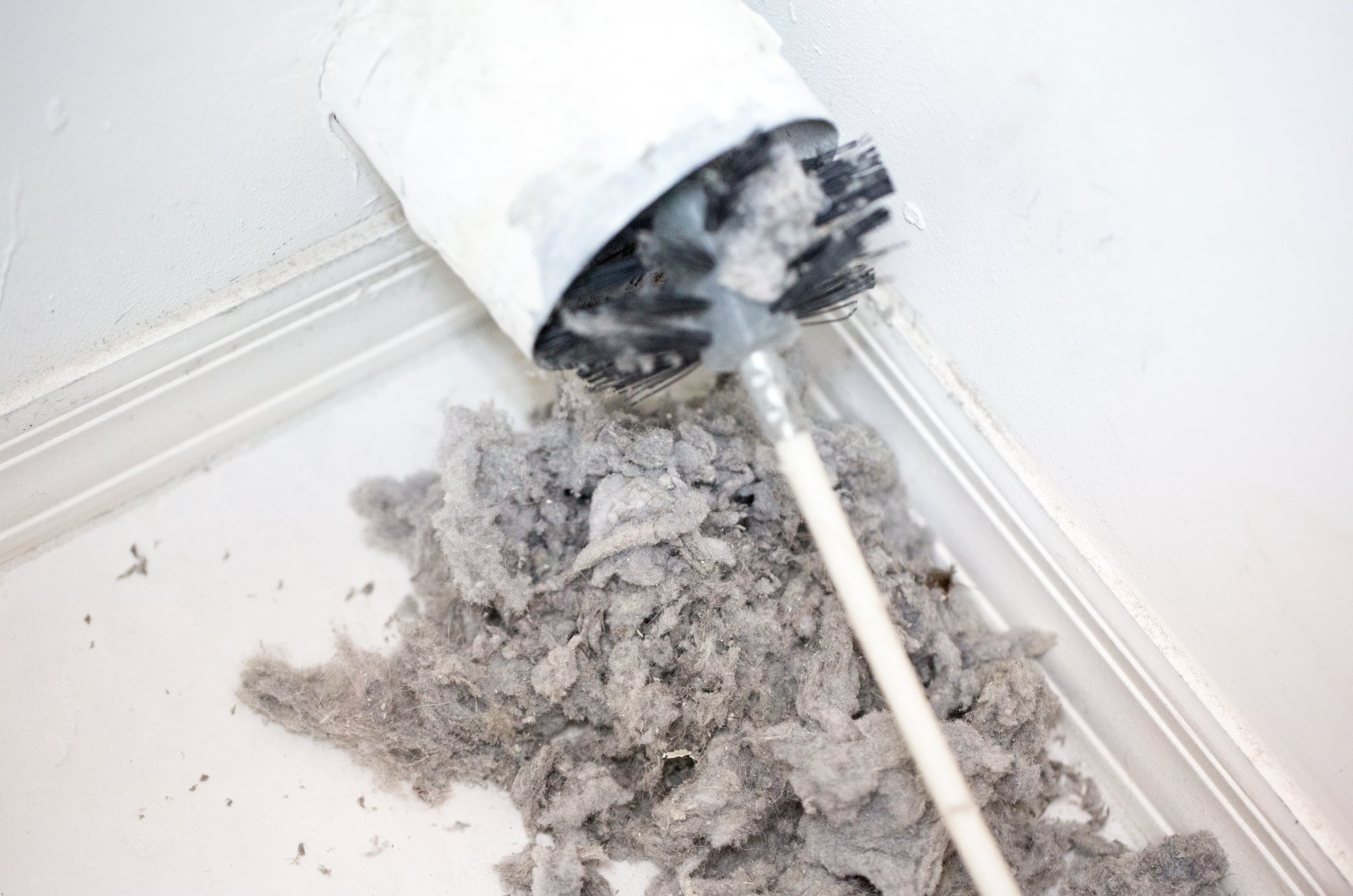 Dryer vent in a home being cleaned out with a round brush. There is a large pile of lint that has been removed from the vent on a white tiled floor. The walls and baseboards are white. The lint is gray. Taken with a Canon 5D Mark 3 camera.  rm